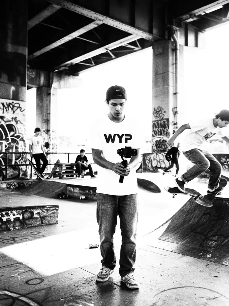 A teenager in a WYP tee filming a friend skateboarding under a bridge